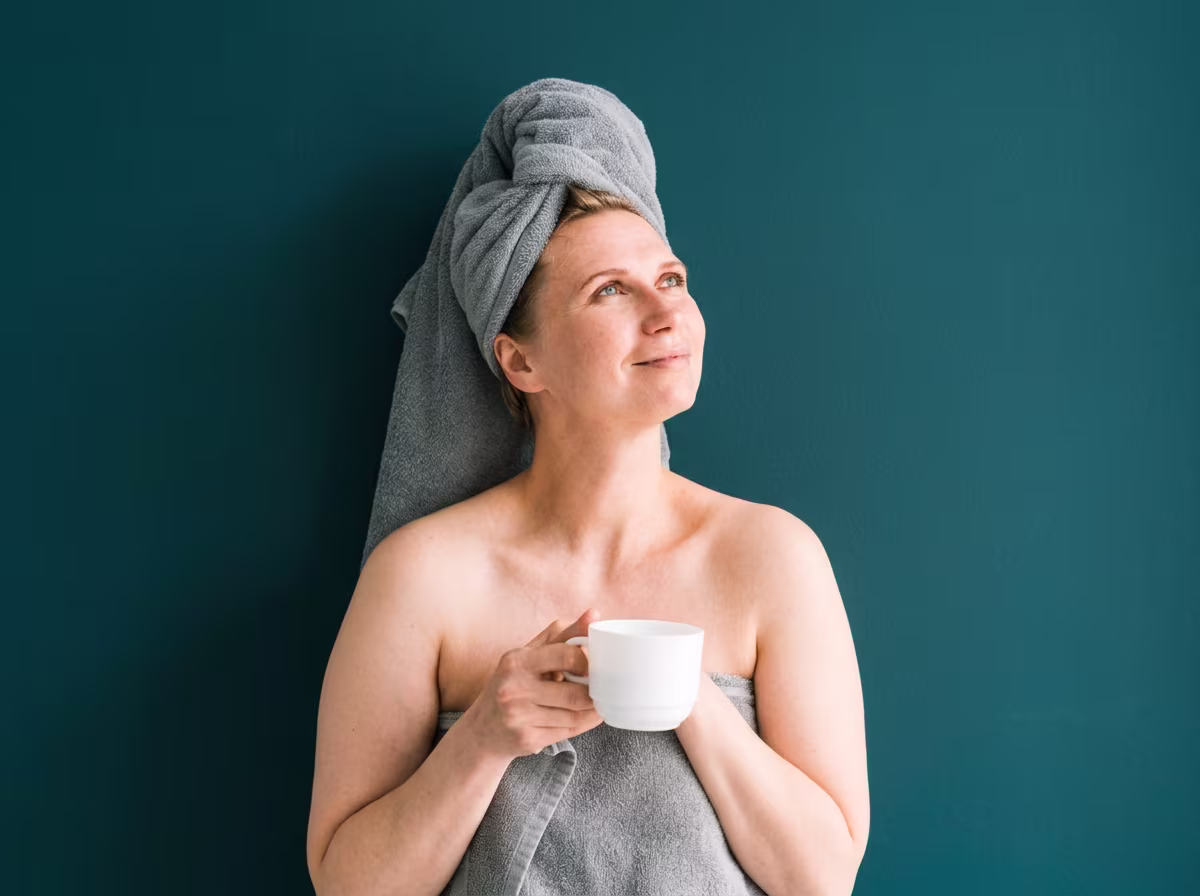 Une femme, enveloppée dans une serviette grise avec une autre serviette enroulée autour de ses cheveux, tient une tasse blanche et regarde vers le haut avec un léger sourire, sur un fond bleu-vert foncé.