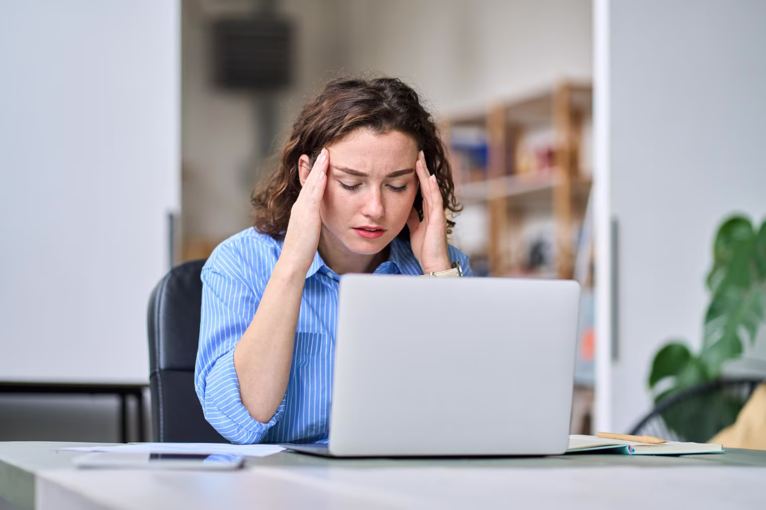 Une jeune femme assise à un bureau devant un ordinateur portable se tient les tempes ; elle affiche une expression tourmentée, semblant stressée ou dépassée.