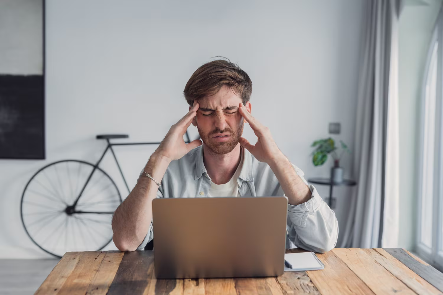 Un homme est assis à une table en bois devant un ordinateur portable ; il se tient la tête à deux mains, les yeux fermés, et semble stressé. La scène se déroule dans une pièce lumineuse, avec un vélo et des plantes en arrière-plan.