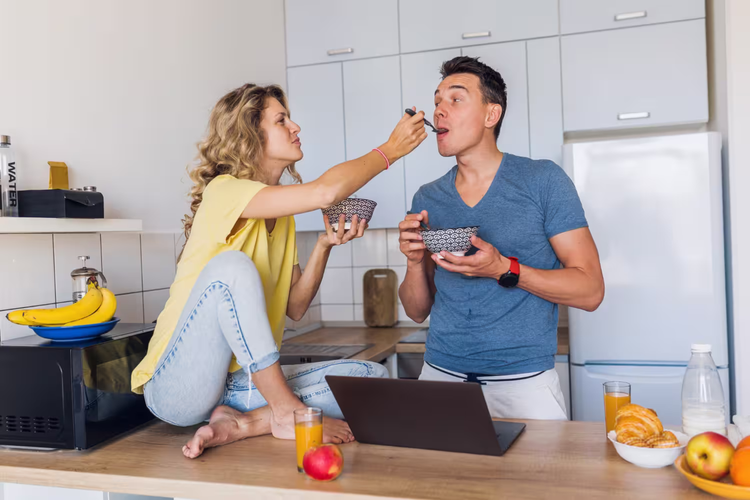 Une femme, assise pieds nus sur un plan de travail de cuisine, donne à manger à un homme à l'aide d'une cuillère ; tous deux tiennent des bols. Sur le plan de travail, dans une cuisine moderne, se trouvent également un ordinateur portable, des fruits, des croissants et des verres de jus d'orange.
