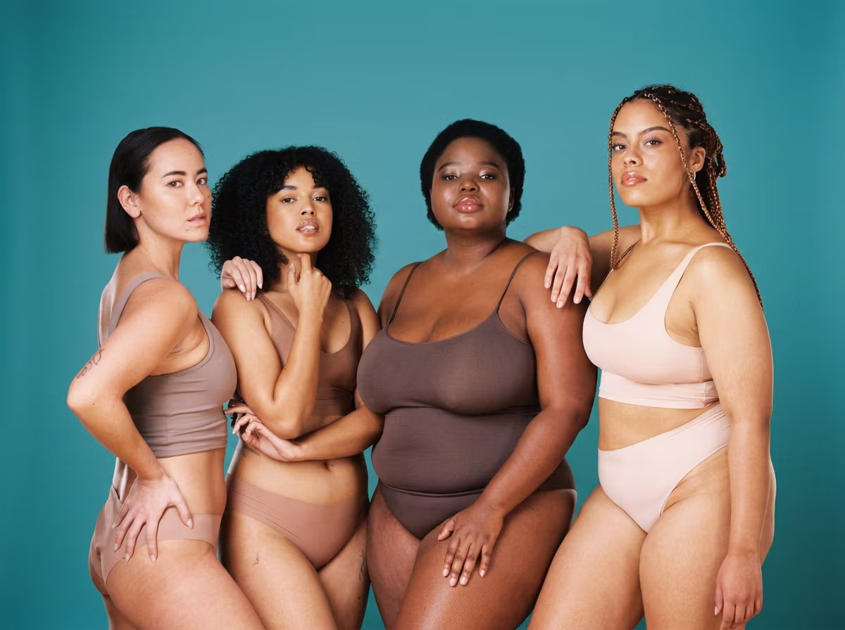 Quatre femmes aux morphologies et origines variées, en sous-vêtements aux tons nude, posant ensemble sur fond bleu canard