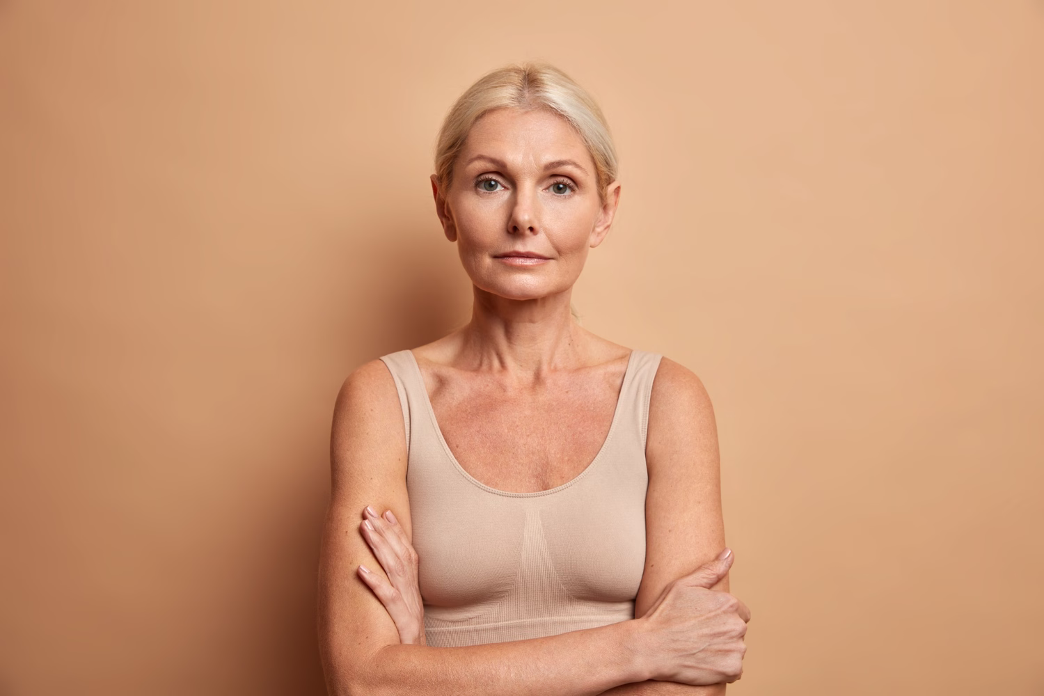 Femme d'une cinquantaine d'années aux cheveux blonds courts, en brassière beige, bras croisés, sur fond beige