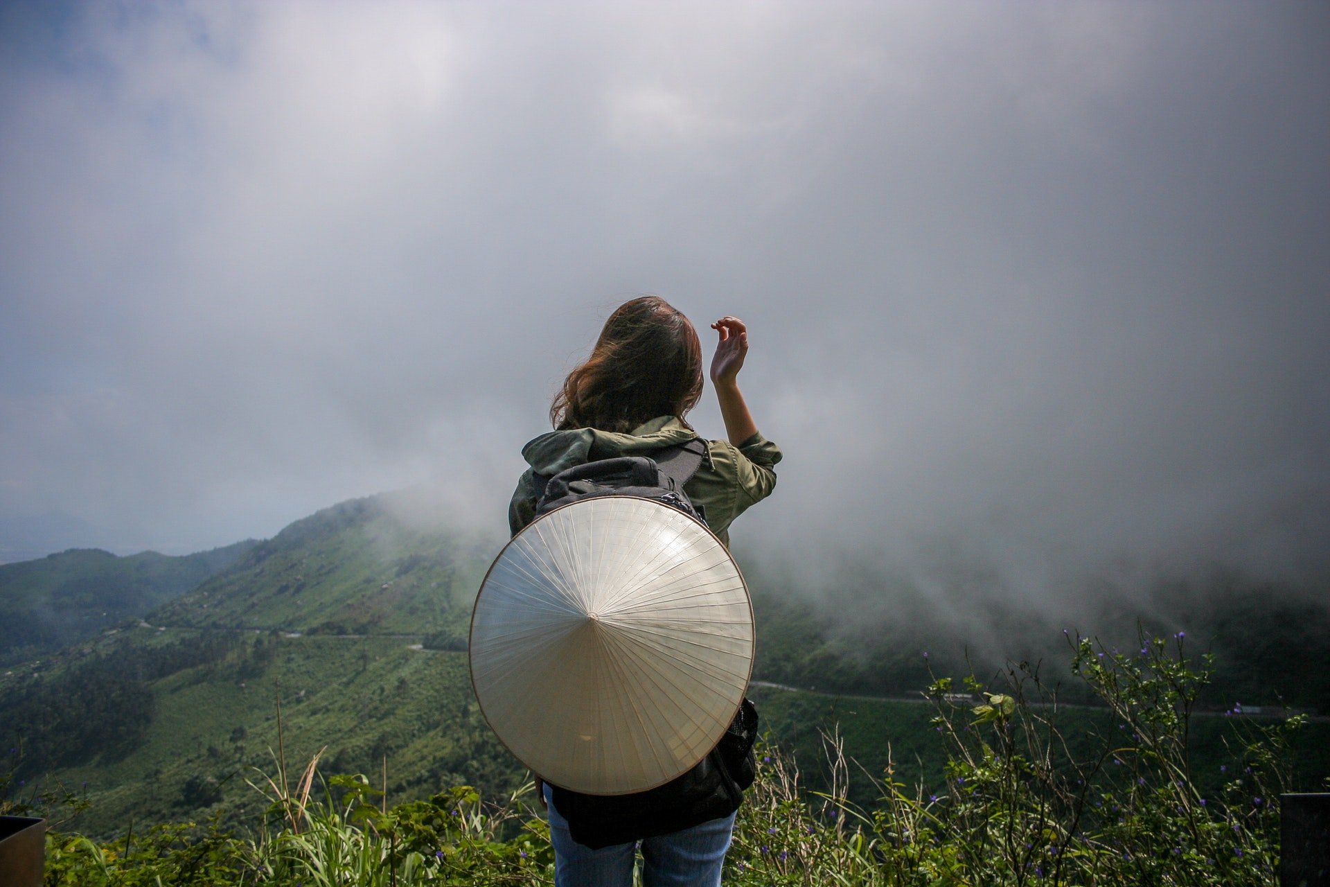 Voyageuse de dos portant un chapeau conique, face à des collines verdoyantes dans la brume